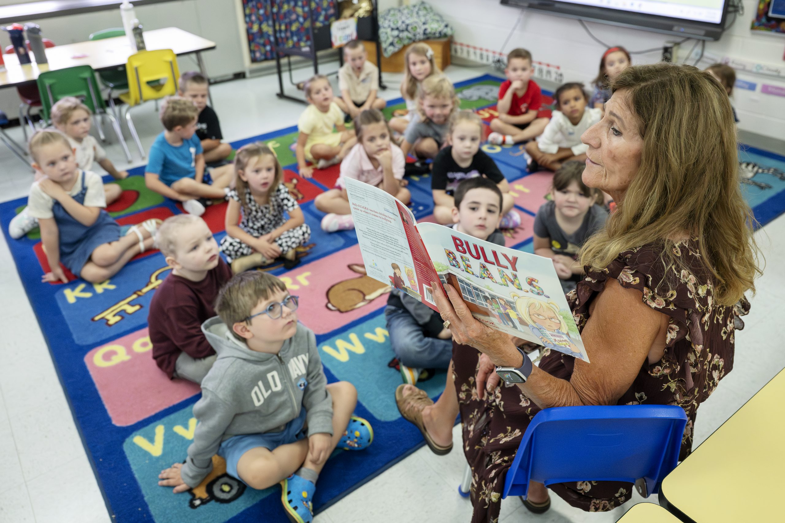 Students sit on a carpet while a teacher reads them a book.